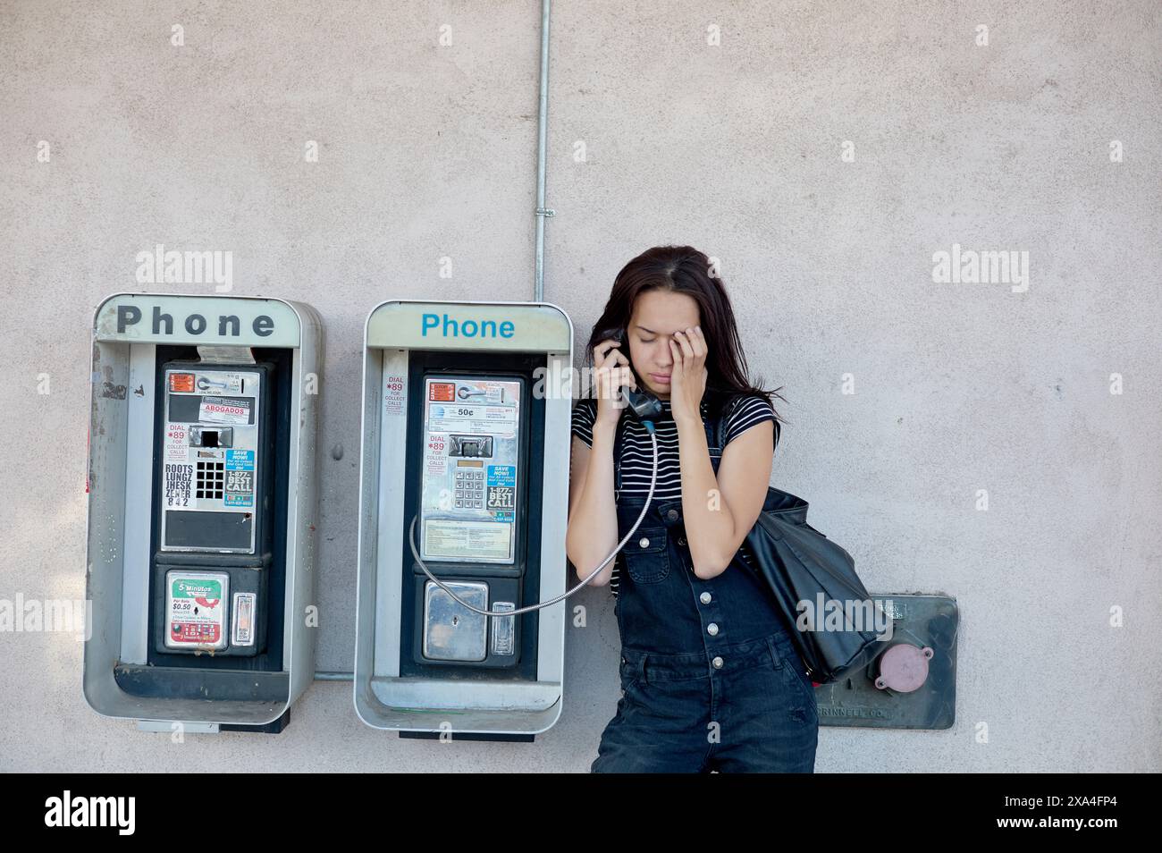 Une femme se tient debout entre deux téléphones publics, engagée dans une conversation sur l'un d'eux, les yeux fermés et un léger froncement de sourcils, indiquant peut-être une discussion sérieuse ou troublante. Banque D'Images