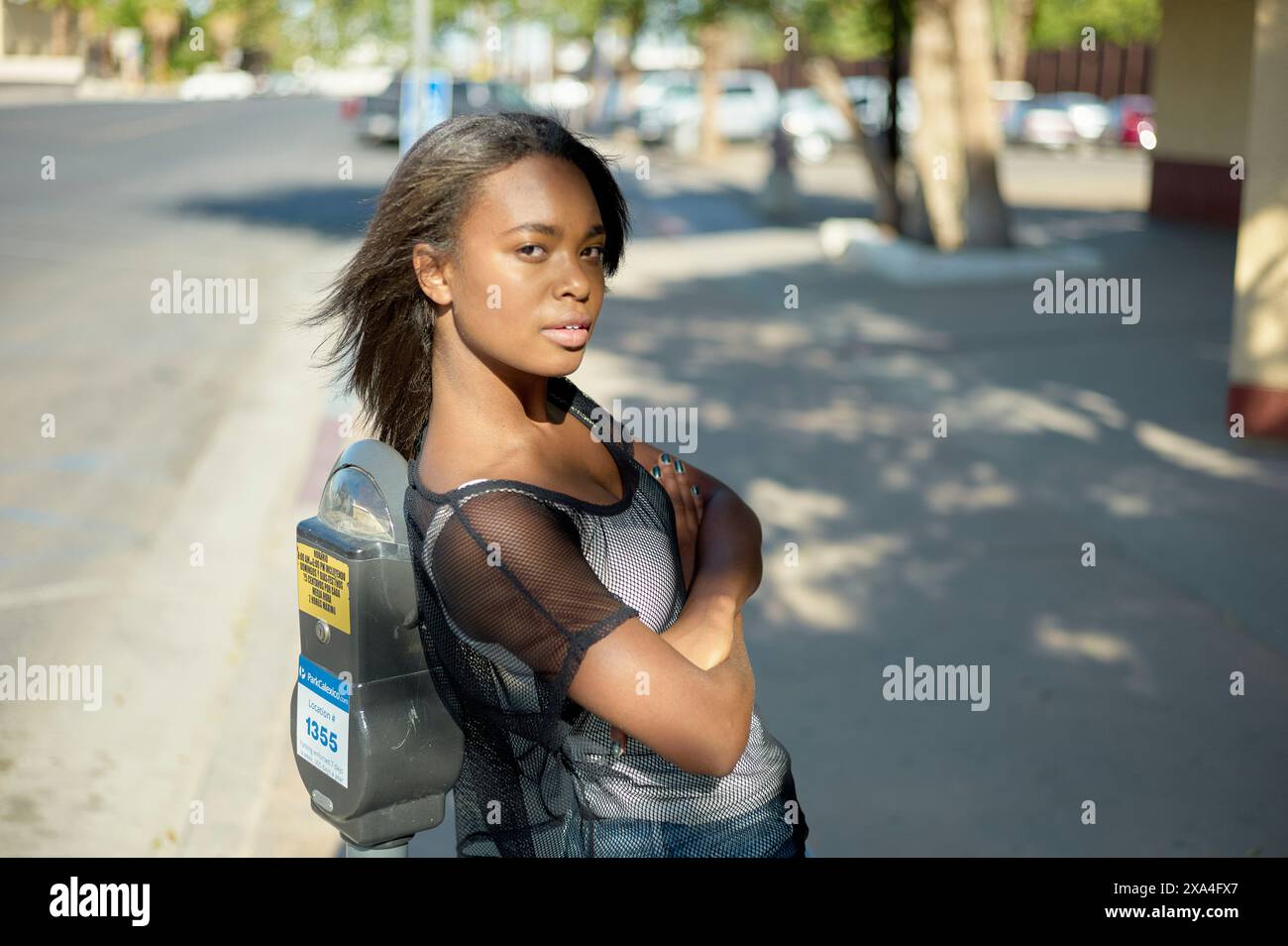 Une femme confiante se tient dans une rue ensoleillée avec les bras croisés, portant un top en maille noire et un sac à dos, regardant par-dessus son épaule la caméra. Banque D'Images