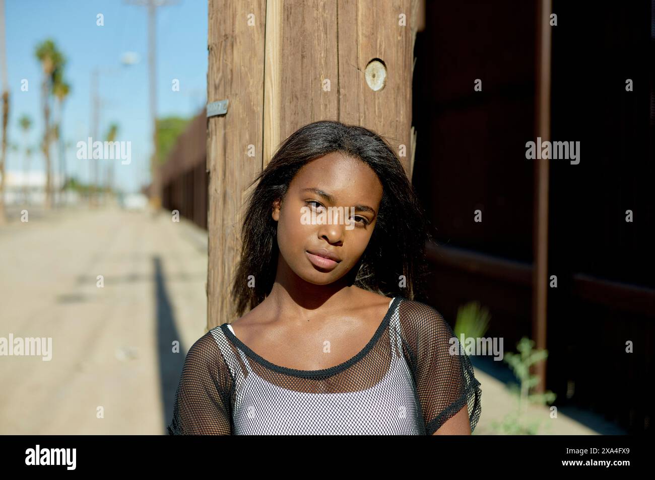 Une femme se tient près d'un poteau en bois dans une rue ensoleillée, portant un top en maille au-dessus d'une tenue décontractée, et regardant l'appareil photo. Banque D'Images
