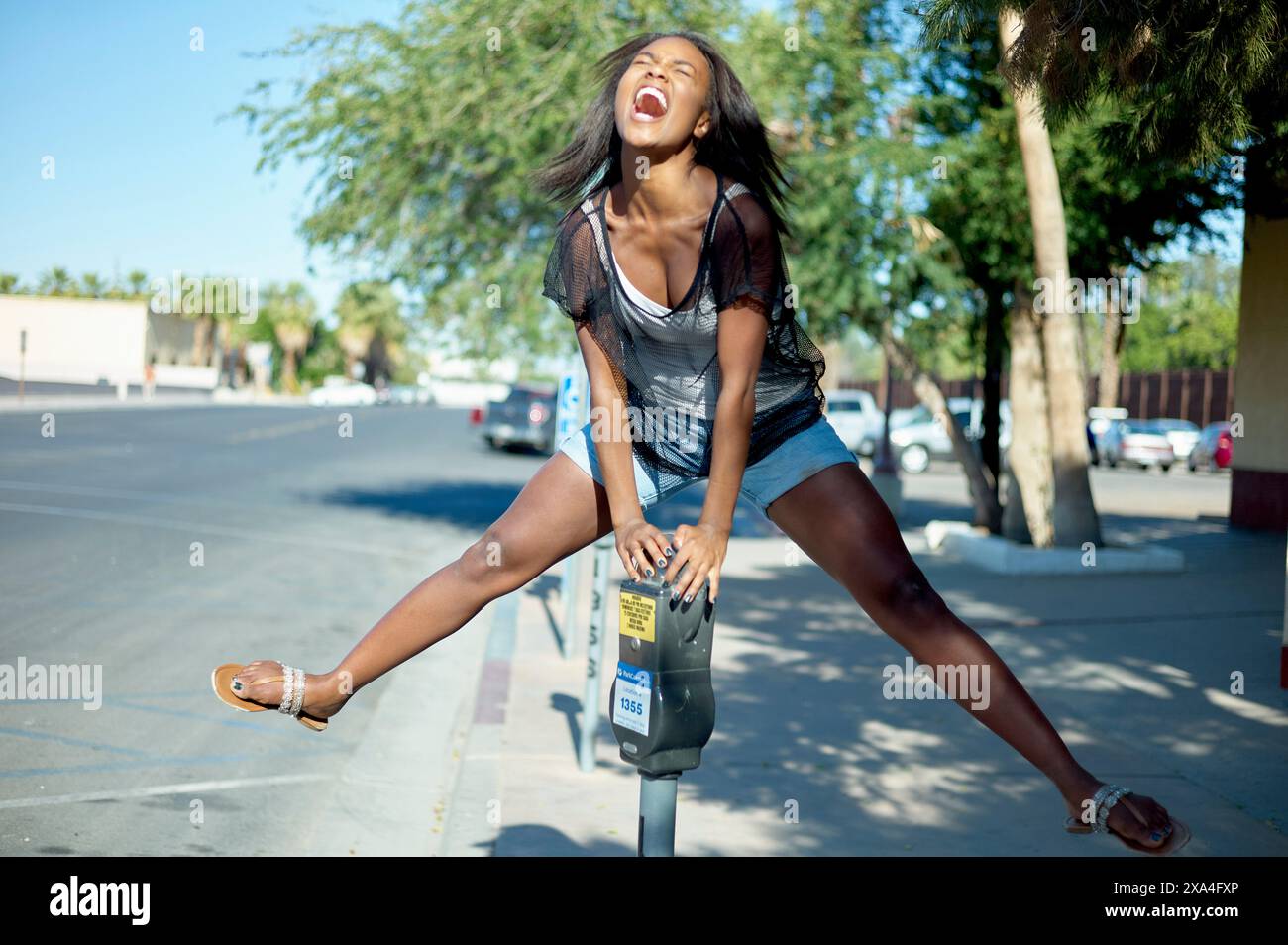 Une jeune femme vêtue d'un top noir transparent et d'un short pose en toute confiance sur un trottoir de la ville avec un pied sur un parcmètre, riant et regardant vers le haut. Banque D'Images