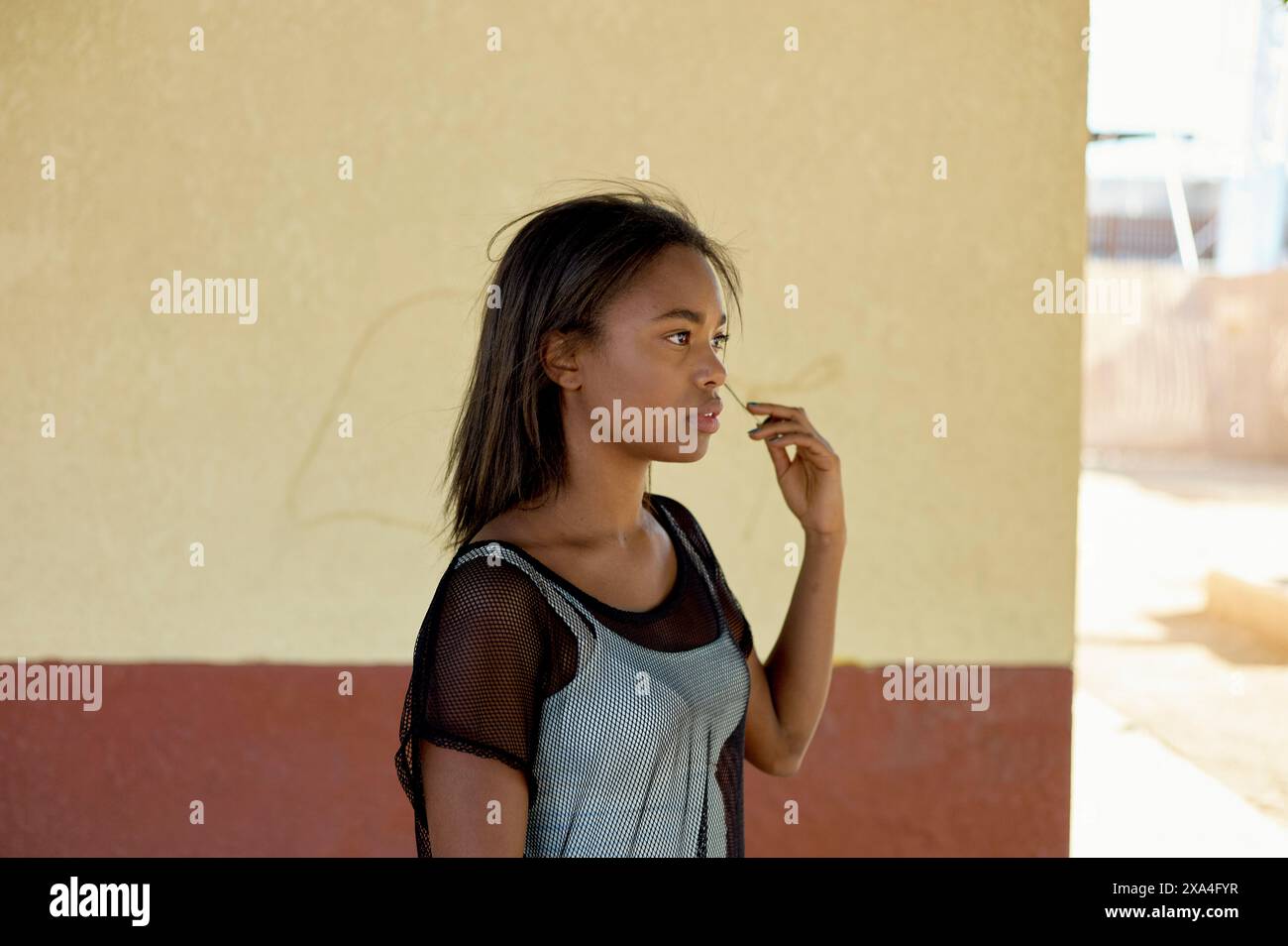 Une jeune fille se tient contre un mur jaune pâle, son regard dirigé sur le côté, les cheveux balayés par le vent. Banque D'Images