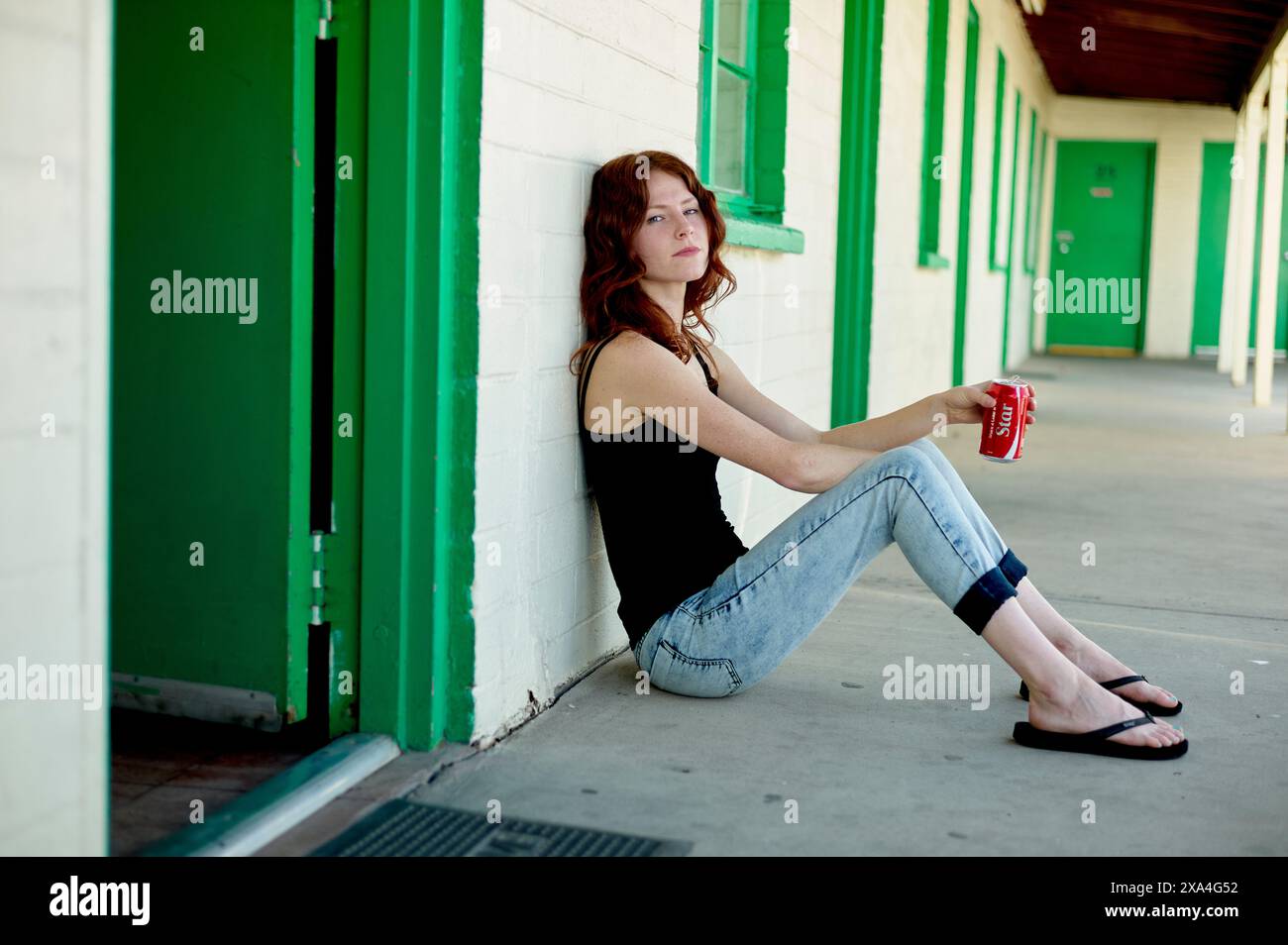 Une jeune femme aux cheveux roux est assise sur le sol, appuyée contre un mur, avec une tasse rouge à la main. Banque D'Images