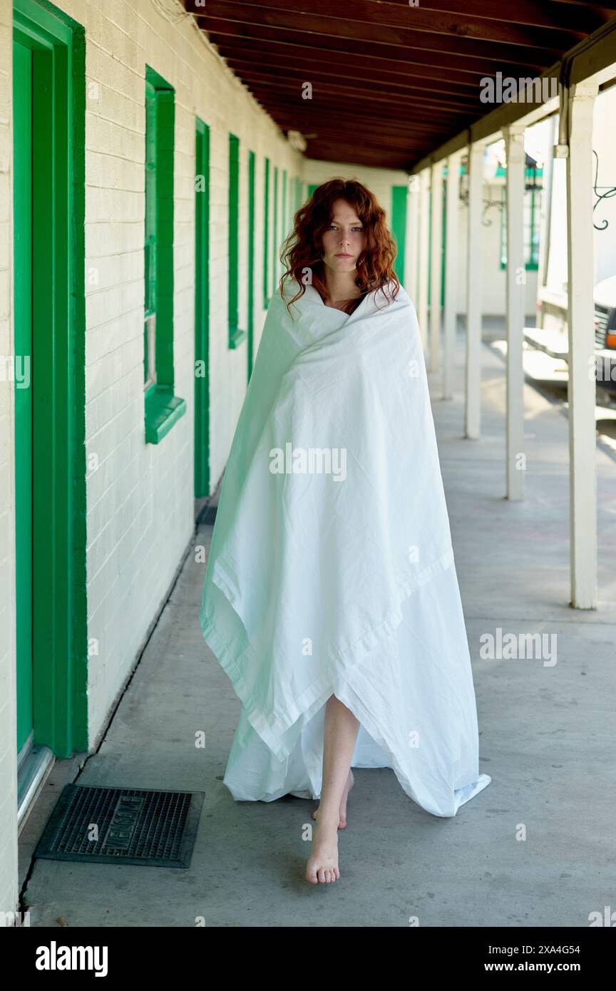 Une femme enveloppée dans un drap blanc marche pieds nus sur un trottoir près d'un bâtiment avec des portes et des fenêtres vertes. Banque D'Images