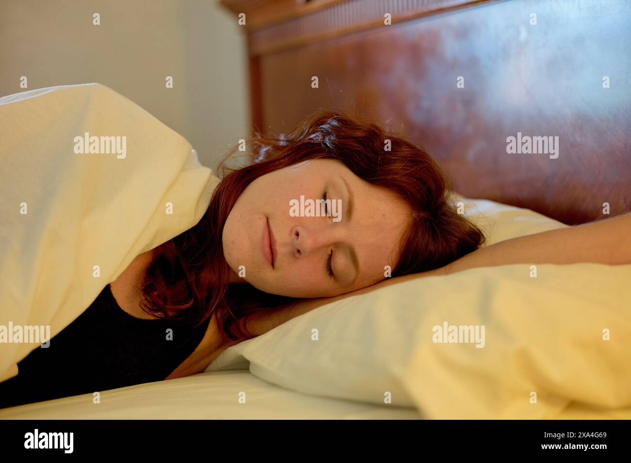 Une femme dort paisiblement dans un lit avec des draps blancs et une tête de lit en bois. Banque D'Images