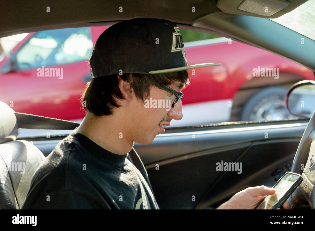 Un jeune homme portant des lunettes et une casquette de baseball est assis dans le siège du conducteur d'une voiture, souriant en regardant un smartphone dans ses mains. Banque D'Images