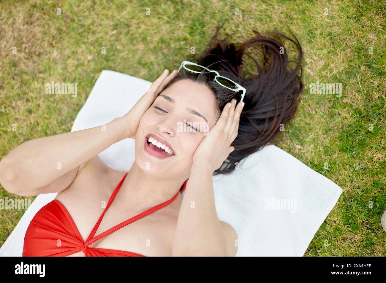 Une femme en maillot de bain rouge est allongée sur une serviette blanche sur l'herbe, riant avec ses mains sur sa tête et ses lunettes de soleil reposant au-dessus de son front. Banque D'Images