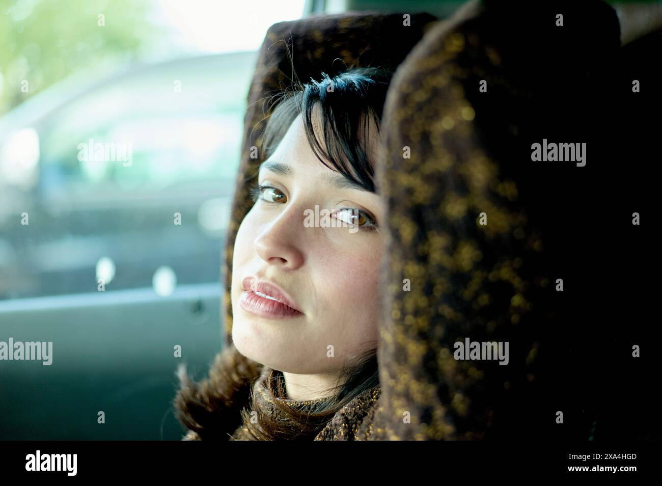 Une femme regarde soigneusement par la fenêtre d'une voiture, ses yeux scintillant contre une lumière naturelle douce, soulignant son expression contemplative. Banque D'Images