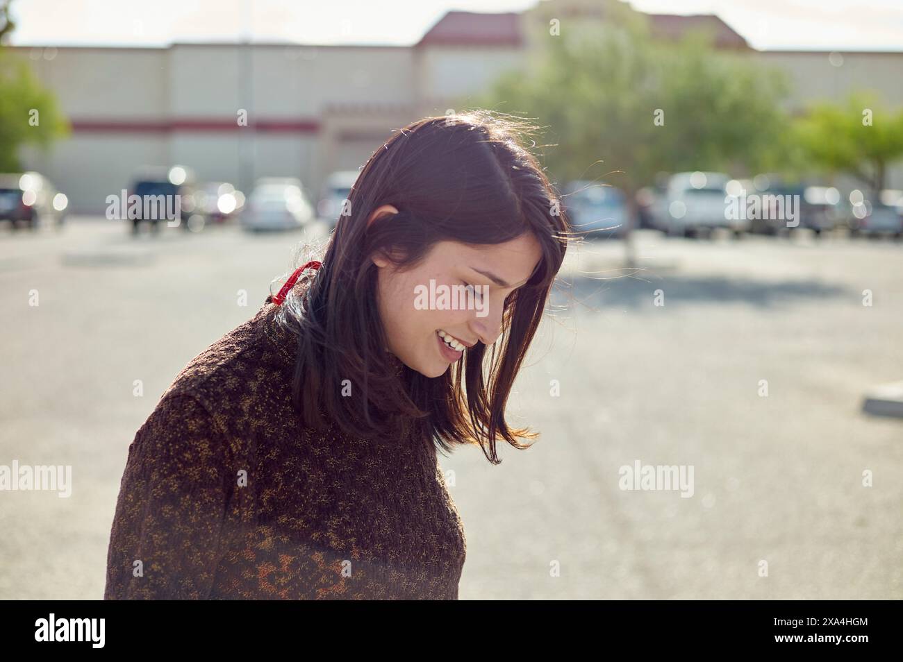 Une jeune femme souriante aux cheveux foncés profitant d'une journée ensoleillée dans un parking, avec des bâtiments visibles en arrière-plan. Banque D'Images