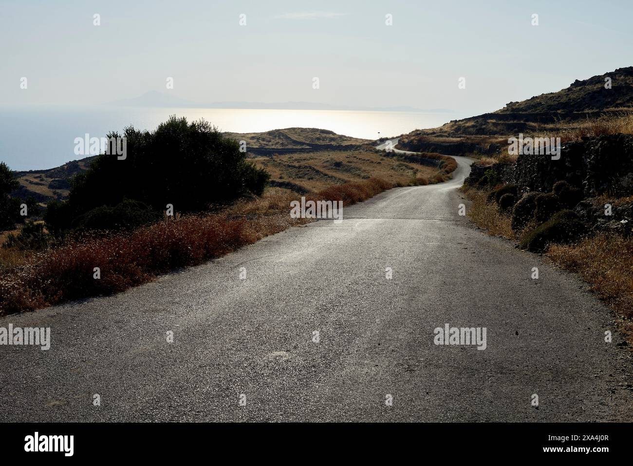 Une route sinueuse descend à travers un paysage sec vers la mer, flanquée d'arbustes et de rochers sous un ciel brumeux. Banque D'Images