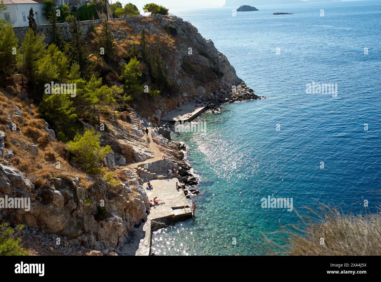 Une vue panoramique sur la côte avec un rivage rocheux, une mer bleu clair, et les gens appréciant une journée ensoleillée près d'une petite jetée. Banque D'Images