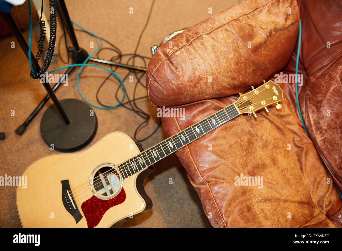 Une guitare acoustique repose sur un canapé en cuir marron avec des câbles étalés sur le sol et un pied de microphone en arrière-plan. Banque D'Images