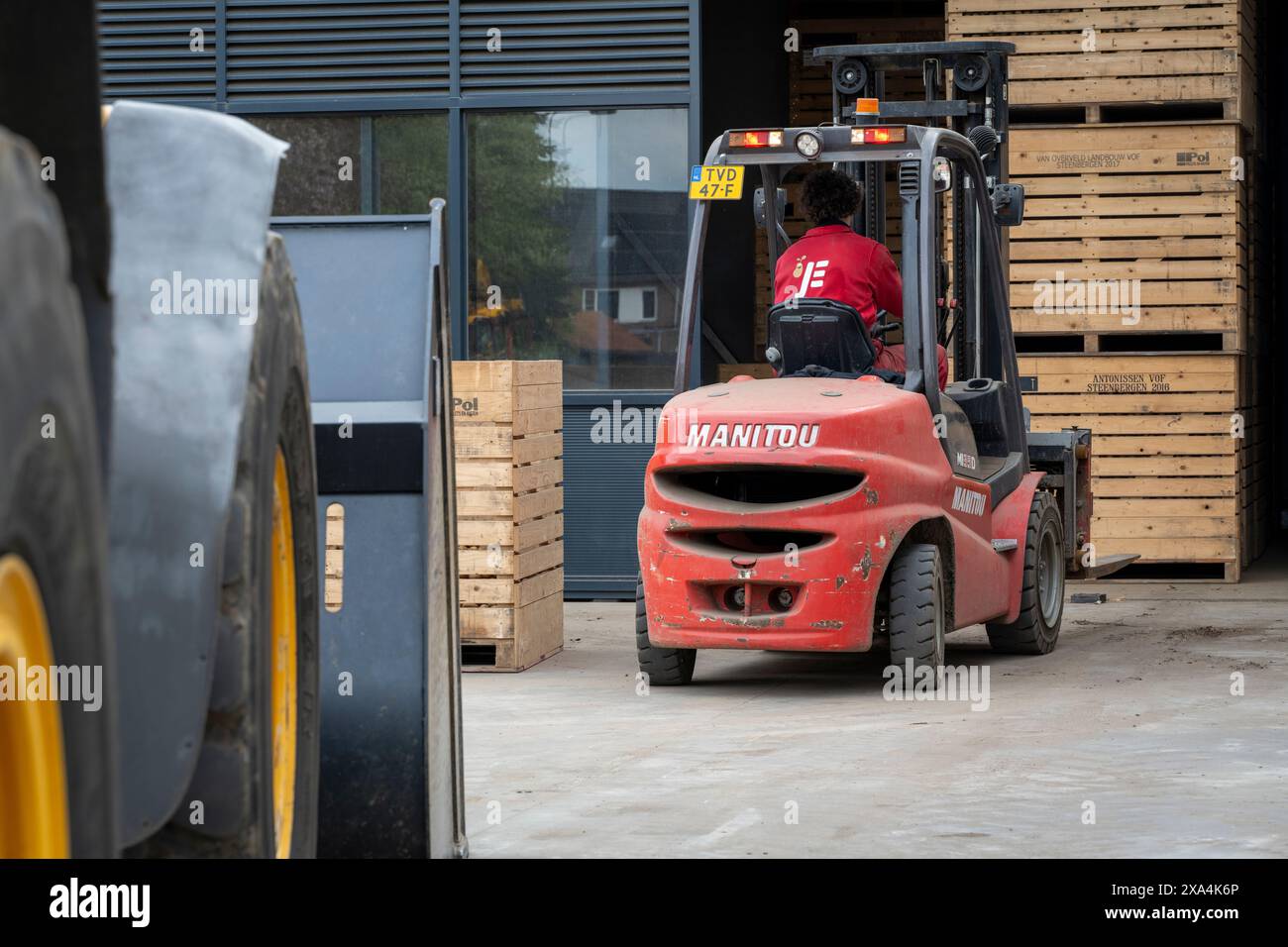 Un travailleur exploite un chariot élévateur rouge Manitou à l'extérieur entre un entrepôt et des palettes en bois empilées. Banque D'Images