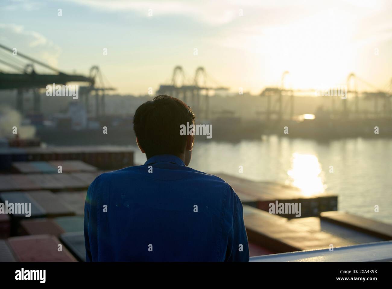 Un homme est vu de derrière regardant un port maritime occupé avec des grues et des conteneurs de fret au lever ou au coucher du soleil. Banque D'Images