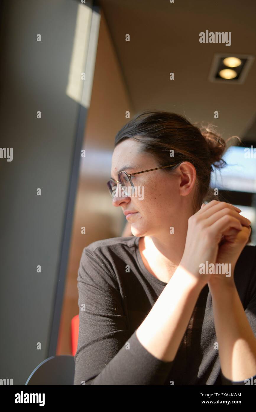 Une femme réfléchie portant des lunettes est assise à l'intérieur avec son menton reposant sur sa main, regardant sur le côté, éclairée par la lumière naturelle. Banque D'Images
