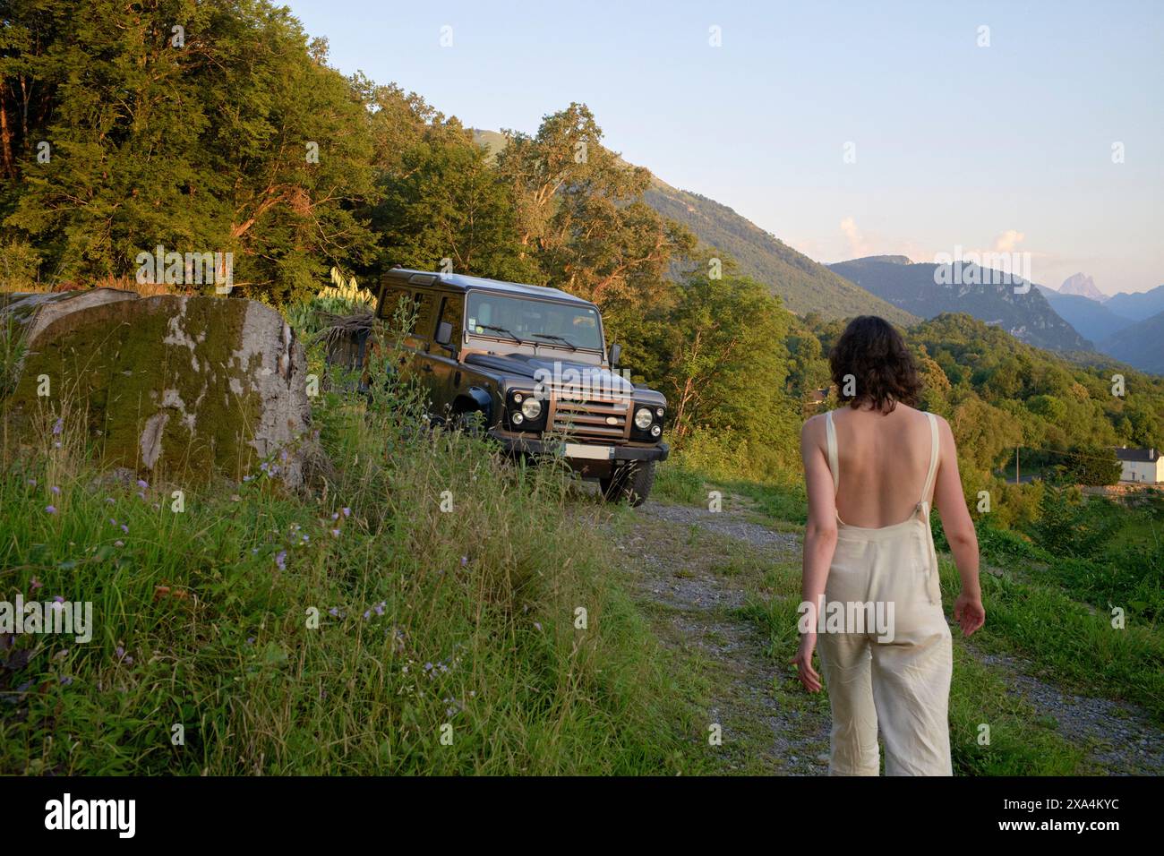 Une personne marchant vers un véhicule hors route garé à côté d'une route de campagne, avec une végétation luxuriante et des montagnes en arrière-plan au crépuscule. Banque D'Images