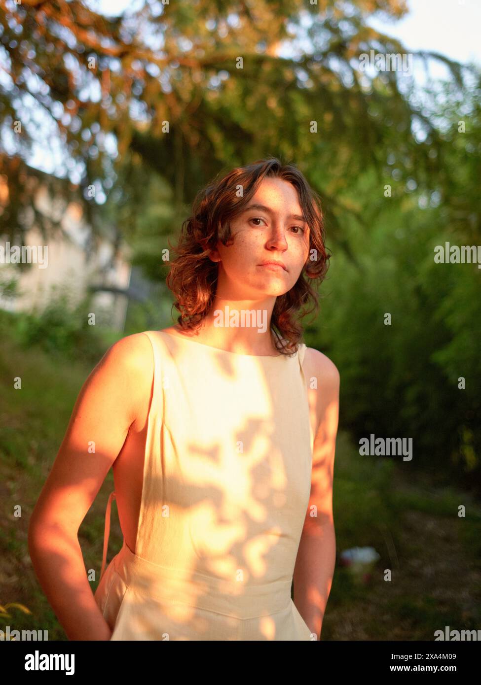 Une jeune femme se tient dehors baignée dans la lueur chaude de la lumière du soleil, avec l'ombre tapée de feuilles projetée sur elle et une toile de fond de verdure luxuriante. Banque D'Images