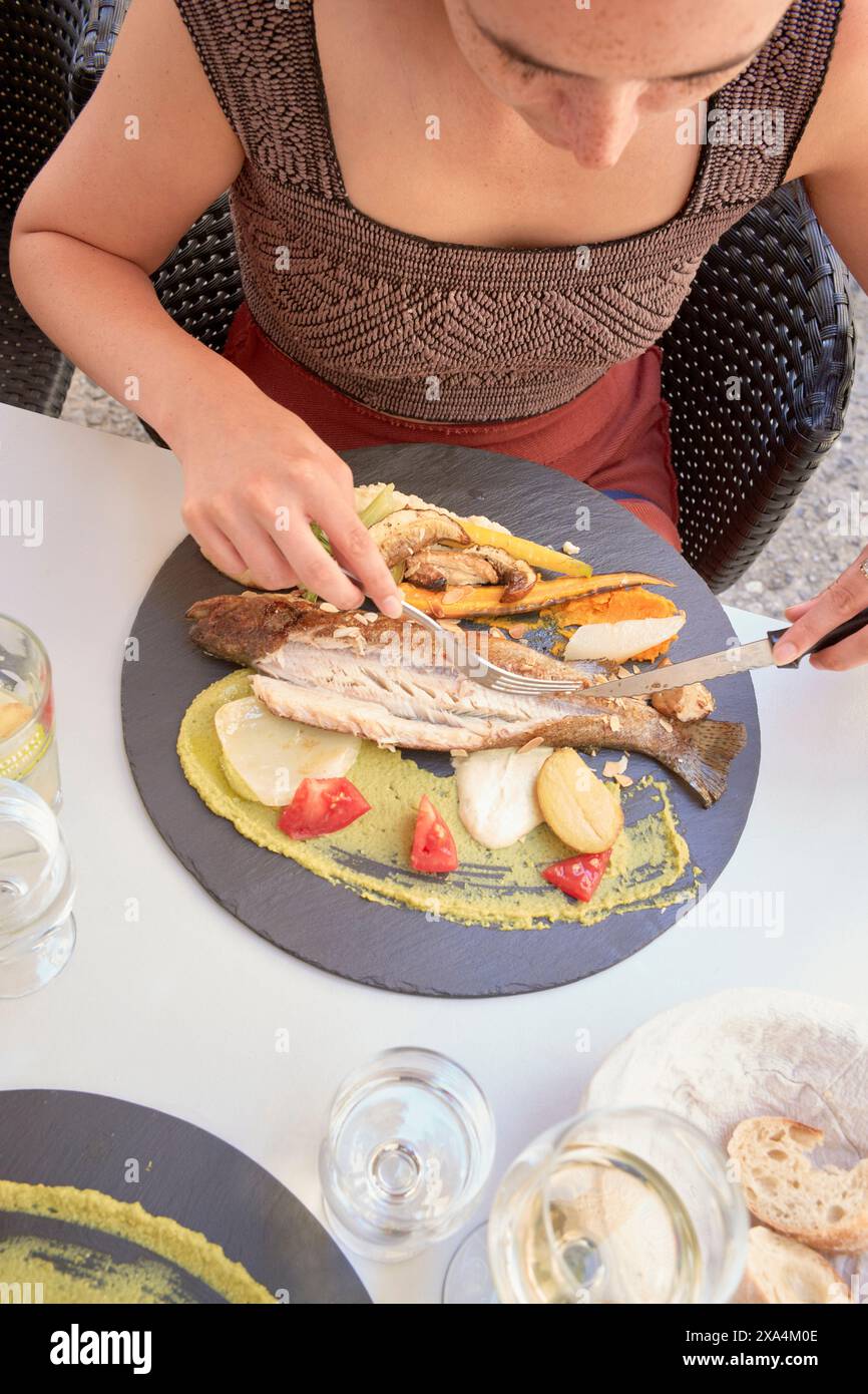 Une personne est assise à une table, concentrée sur couper un poisson grillé servi avec des tranches de citron, des herbes et des légumes sur une assiette ronde en ardoise. Banque D'Images