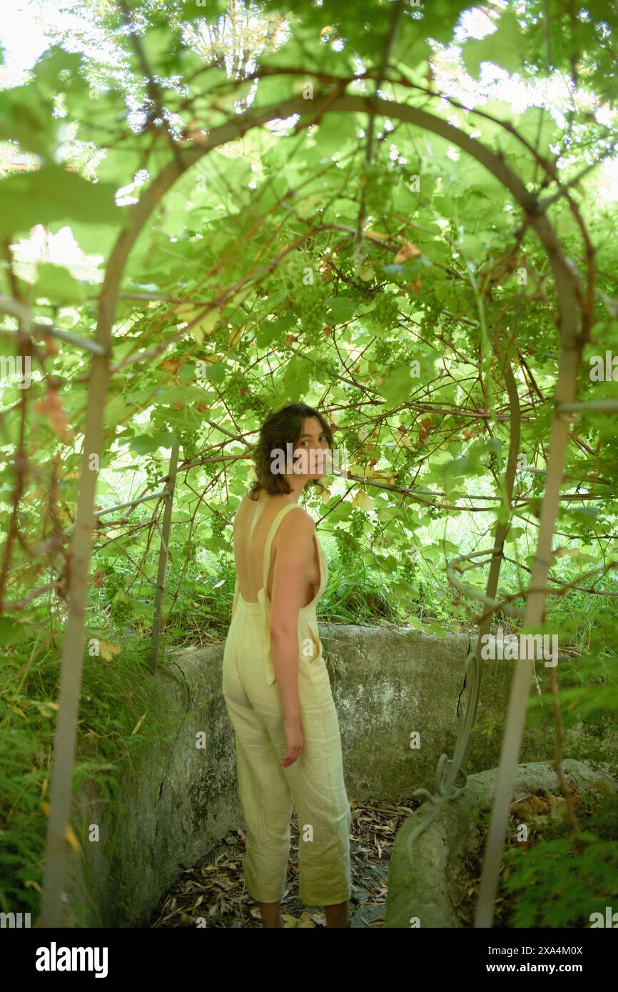 Une femme se tient au milieu de la verdure sous un treillis courbé, tournant la tête pour regarder la caméra avec une expression tranquille. Banque D'Images