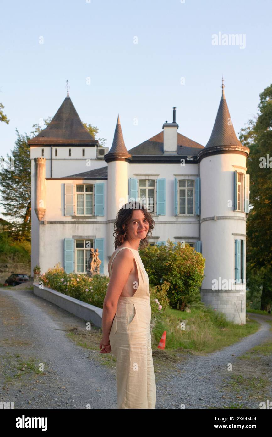 Une femme souriante vêtue d'une tenue crème élégante se dresse devant un élégant château blanc avec des tours jumelles et une végétation luxuriante. Banque D'Images