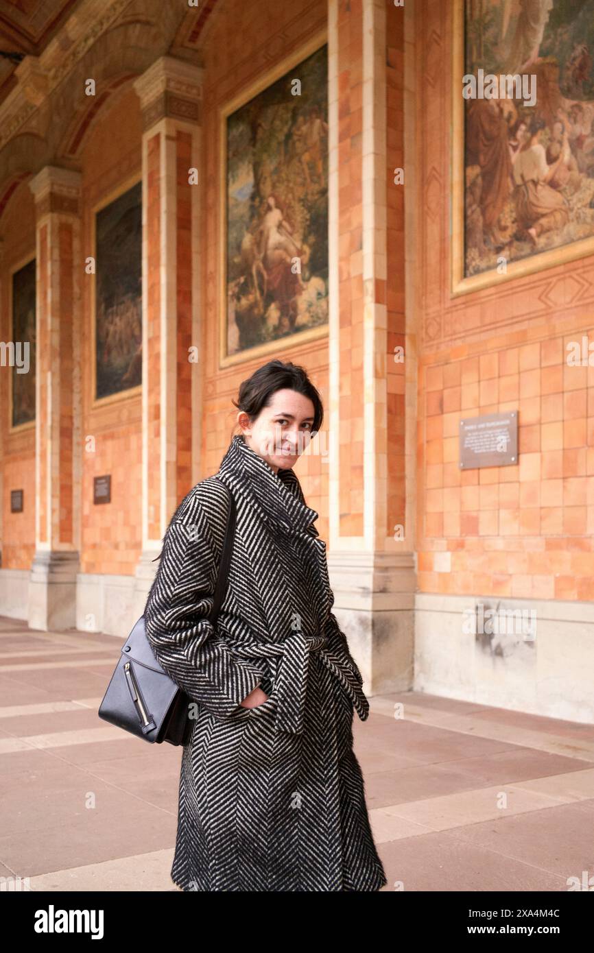 Une femme souriante dans un élégant manteau pied de poule tenant un sac à main se tient devant un bâtiment orné de fresques et d'arches. Banque D'Images