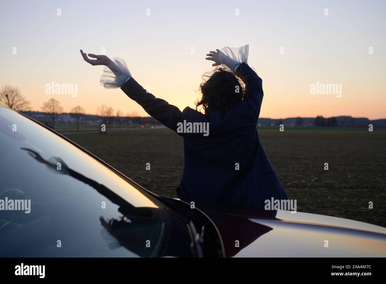 Une personne se tient dos à la caméra, les bras levés d'un geste gracieux, à côté d'une voiture au crépuscule sous un ciel ouvert. Banque D'Images