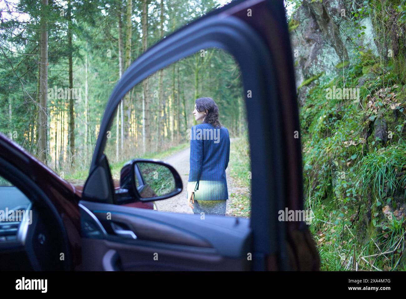 Une jeune femme se tient près d'une route forestière, vue de la porte ouverte d'une voiture avec l'intérieur partiellement visible. Banque D'Images