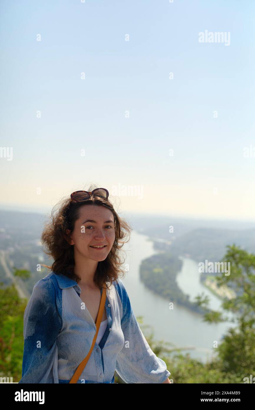 Une femme souriante avec des lunettes de soleil sur la tête pose devant une vue panoramique sur une rivière sinueuse et une verdure luxuriante sous un ciel bleu clair. Banque D'Images