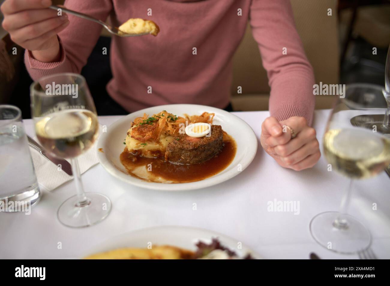 Une personne assise à une table à manger est sur le point de déguster une assiette de steak avec de la sauce, surmontée d'une garniture d'herbes et d'une demi-tranche d'un œuf dur, à côté d'un verre de vin blanc et de divers plats d'accompagnement. Banque D'Images