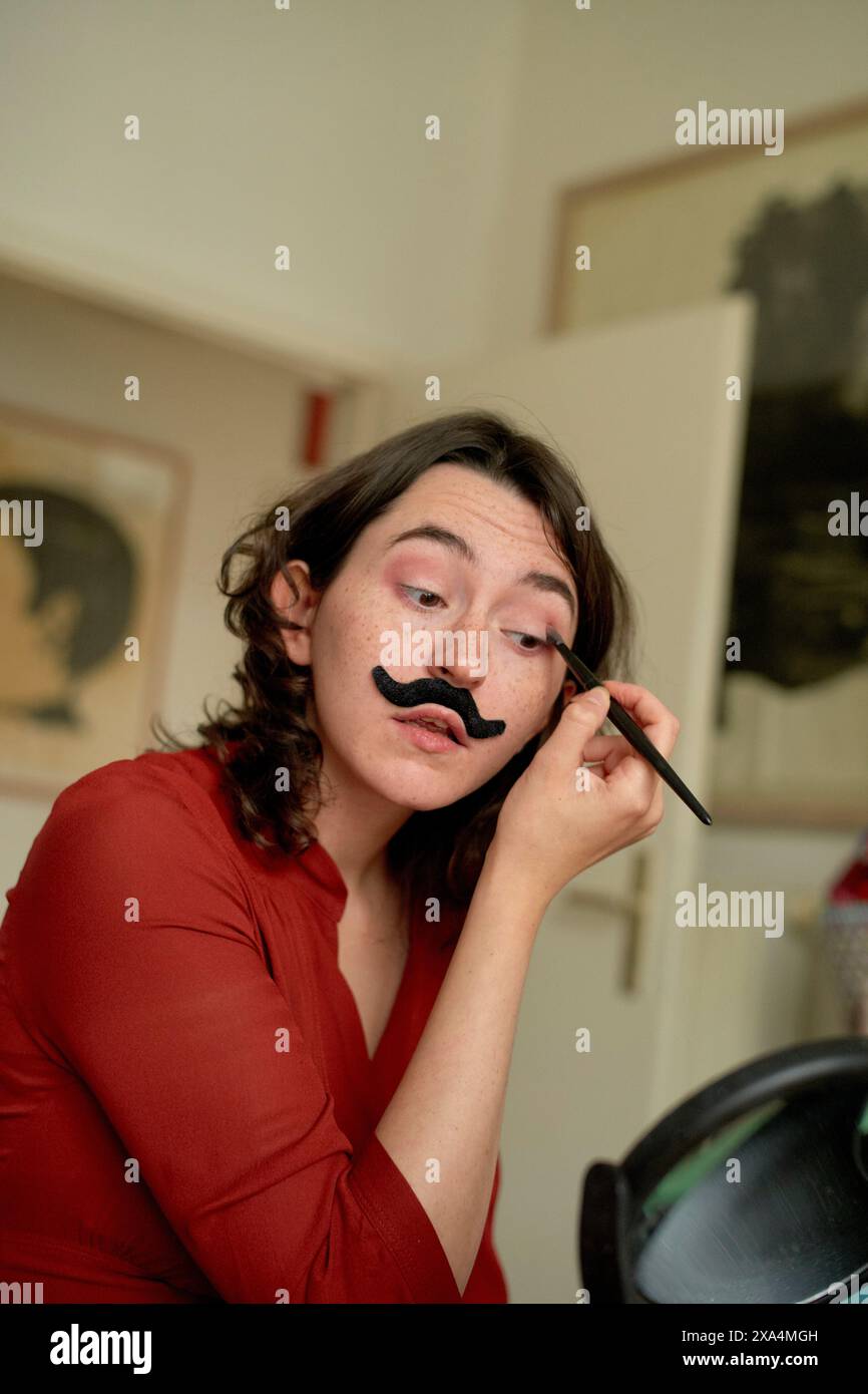 Une femme dans un haut rouge se maquille sur une fausse moustache sur son visage, regardant dans un miroir avec une peinture en arrière-plan. Banque D'Images