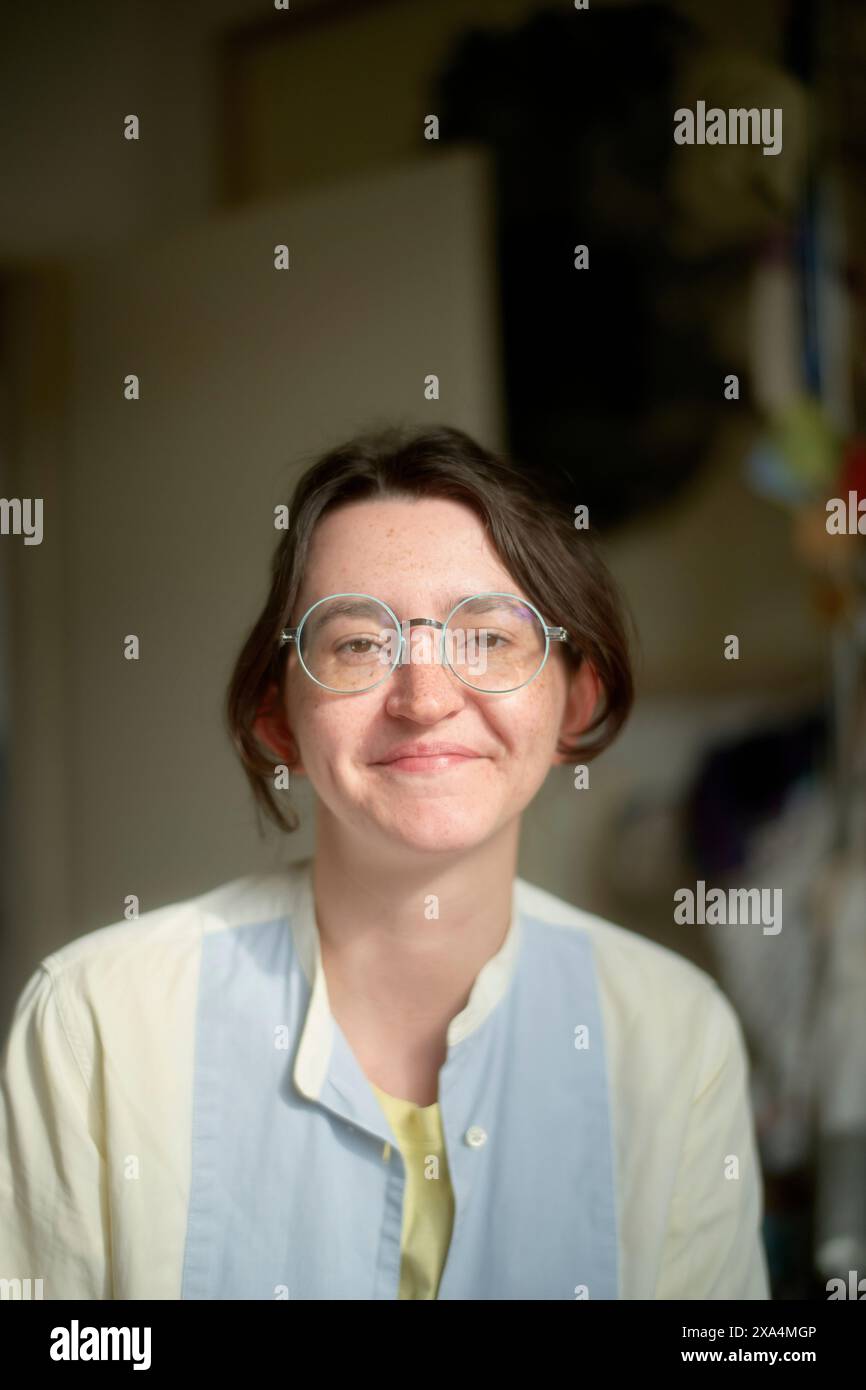 Une jeune femme souriante aux cheveux courts portant des lunettes rondes et une chemise bleu clair sur un haut jaune se tient debout à l'intérieur, avec un fond flou suggérant une pièce avec des objets. Banque D'Images