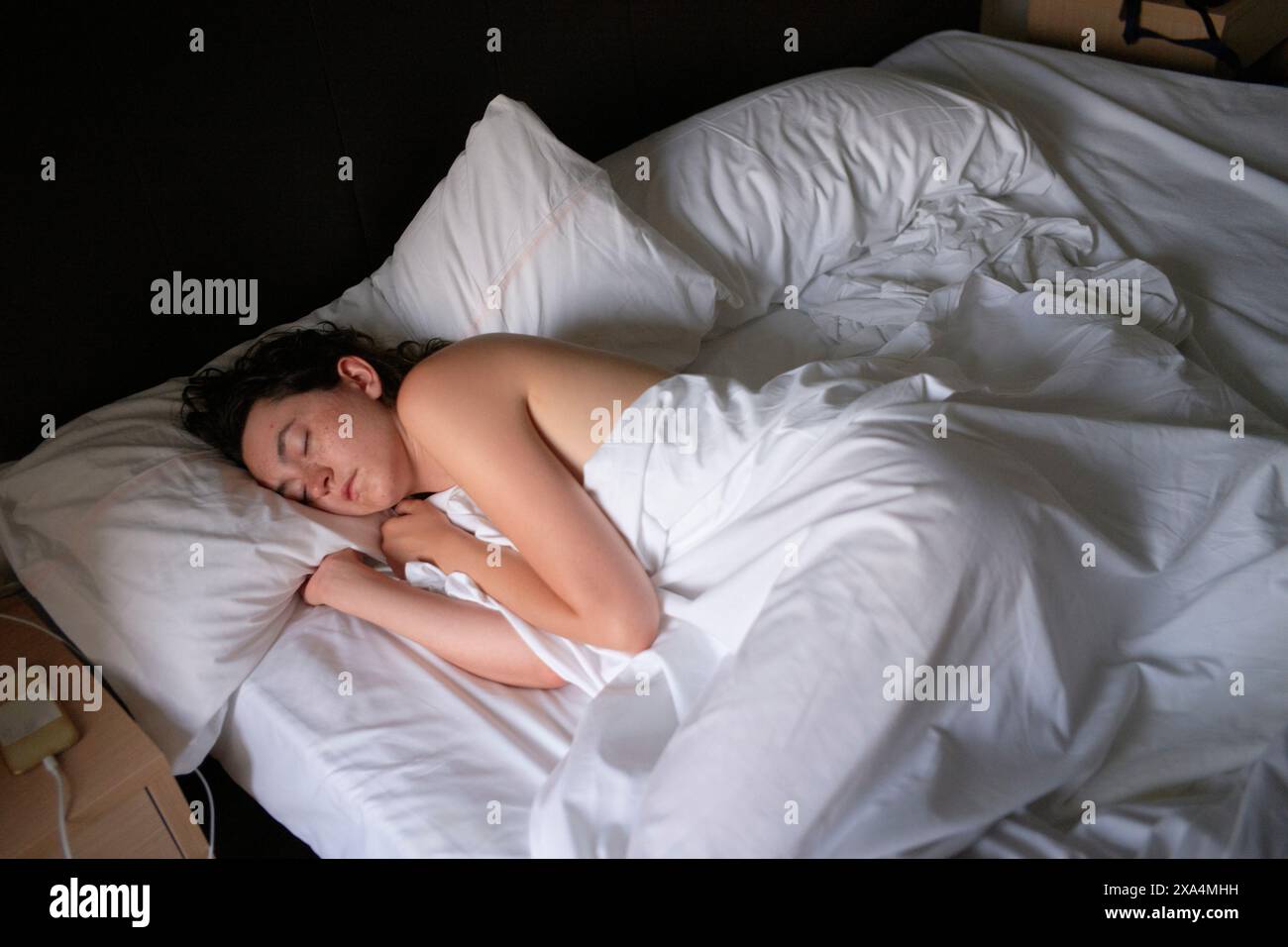 Une jeune femme dort paisiblement sur un lit avec des draps blancs, partiellement recouvert d'une couverture blanche dans une pièce faiblement éclairée. Banque D'Images