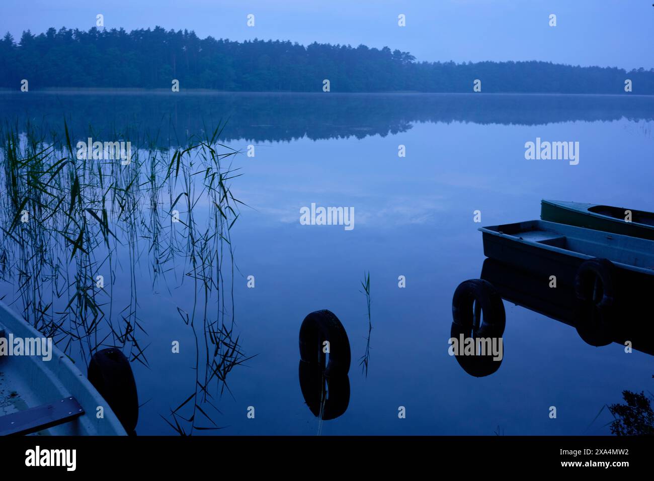 Une scène tôt le matin à un lac calme avec un reflet clair des arbres à la surface de l'eau et des bateaux au bord. Banque D'Images