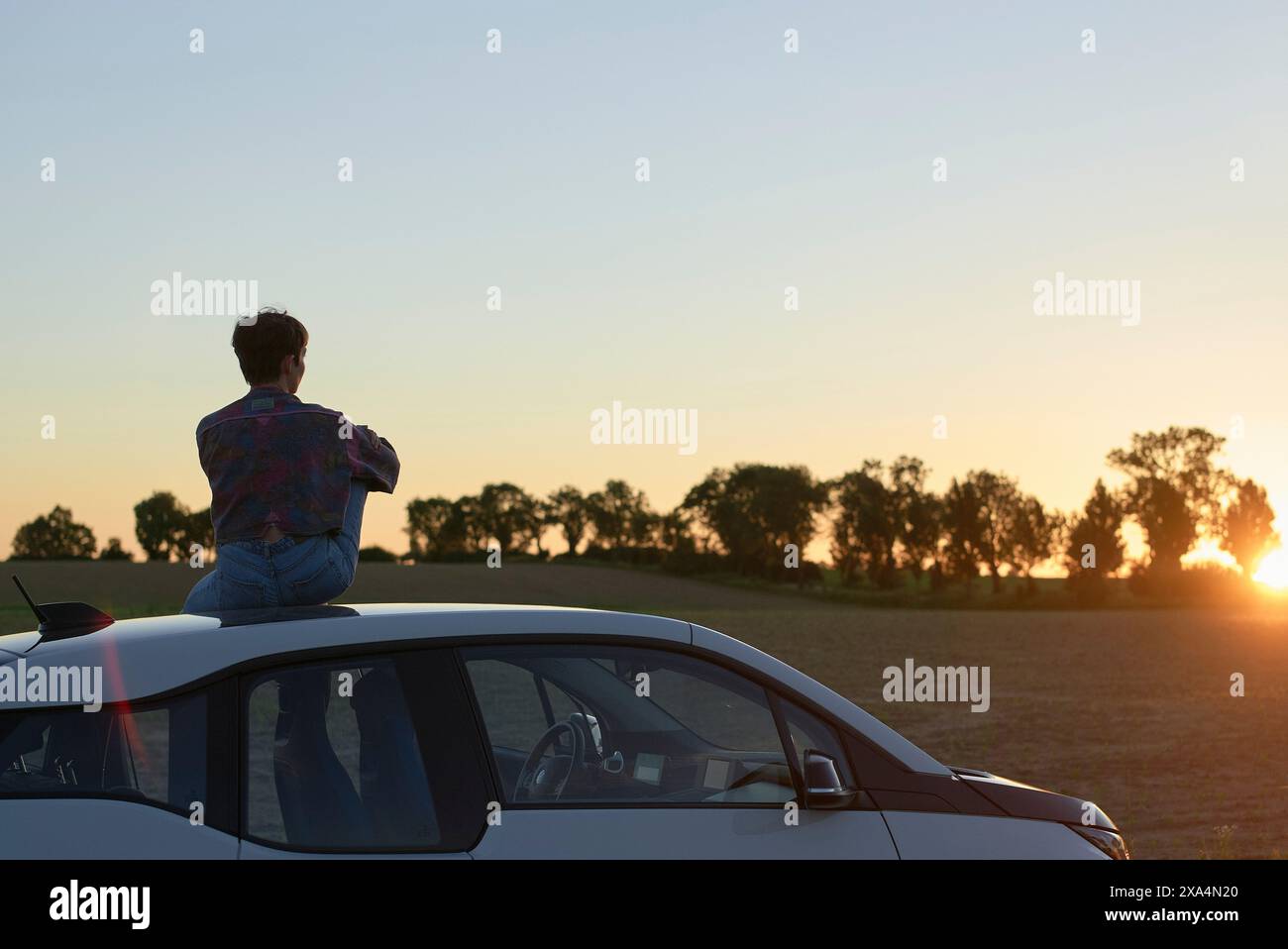 Une jeune femme est assise sur une voiture, regardant un coucher de soleil dans un paysage de campagne. Banque D'Images