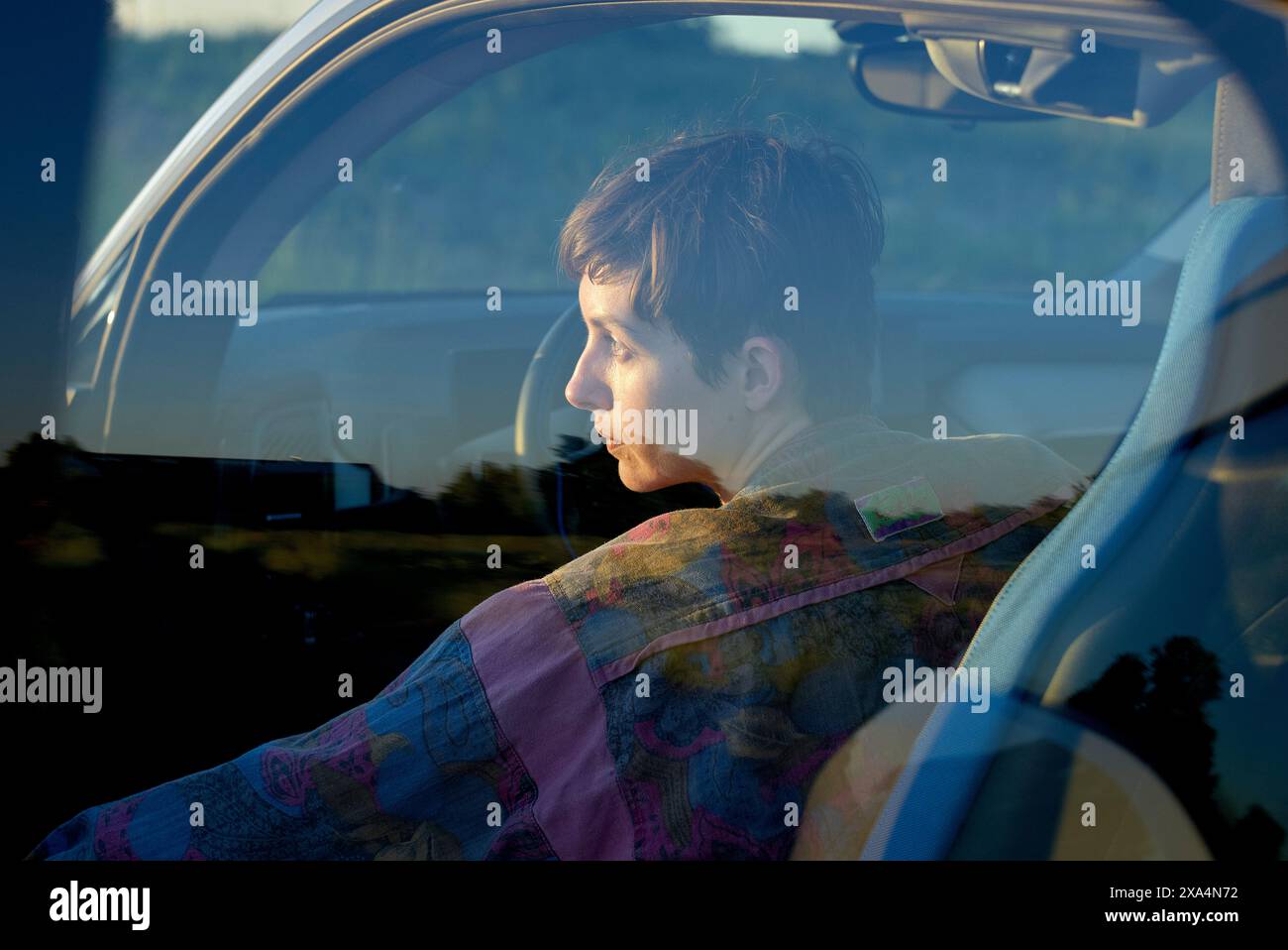 Une jeune femme est assise dans le siège du conducteur d'une voiture, avec une expression réfléchie, regardant par la fenêtre pendant le coucher du soleil. Banque D'Images