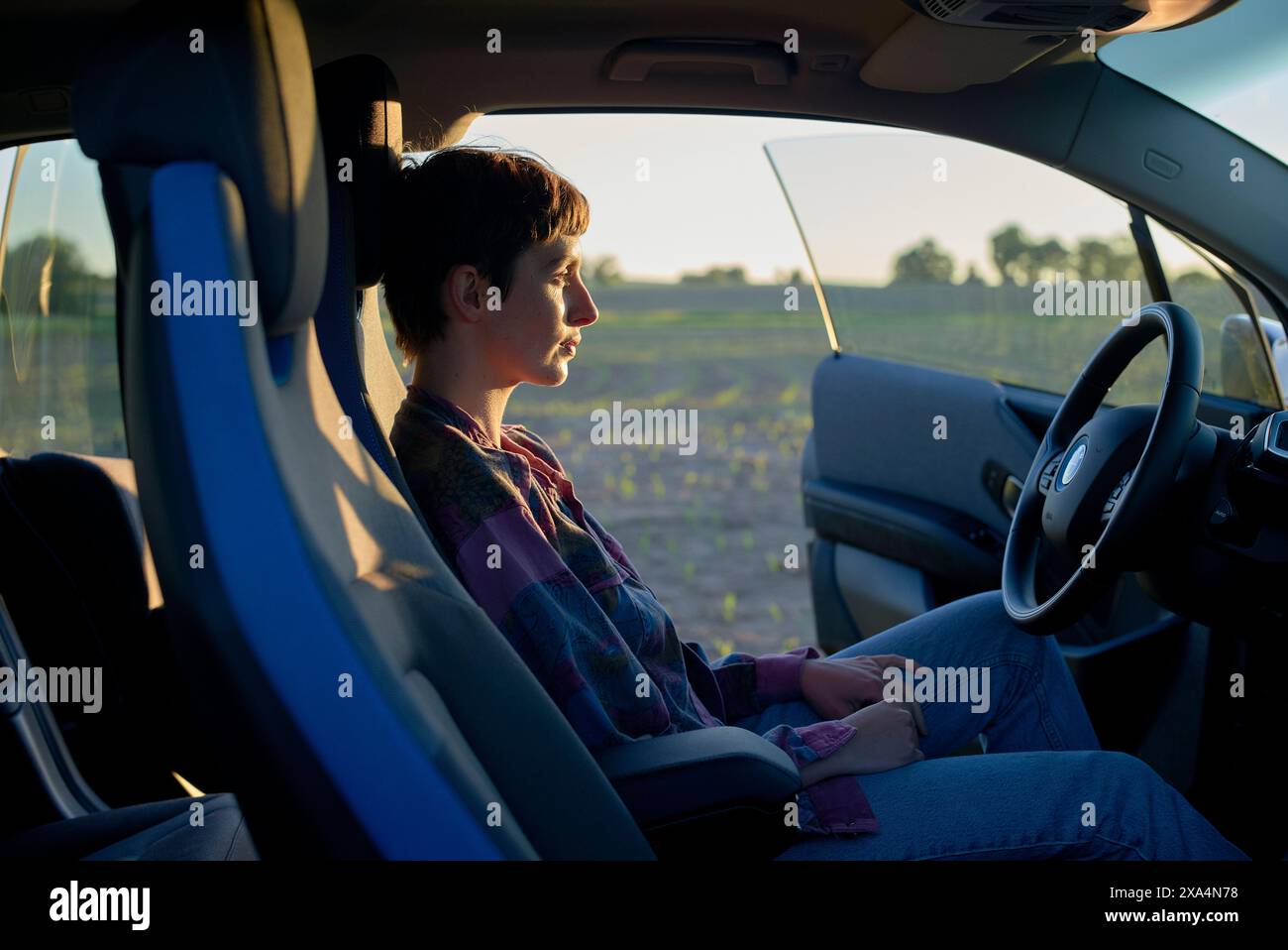 Une jeune femme est assise dans le siège du conducteur d'une voiture au coucher du soleil, regardant soigneusement sur le côté avec une expression calme. Banque D'Images