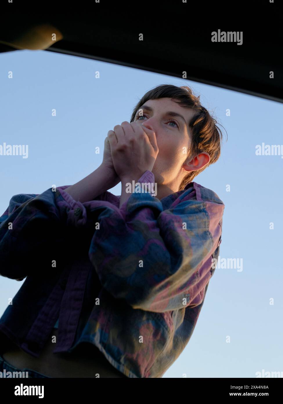 Une jeune femme regarde vers le haut dans le ciel à travers ce qui semble être une fenêtre ouverte, tenant ses mains près de sa bouche dans une pose réfléchie ou anticipée, éclairée par la lumière naturelle sur fond de ciel clair. Banque D'Images