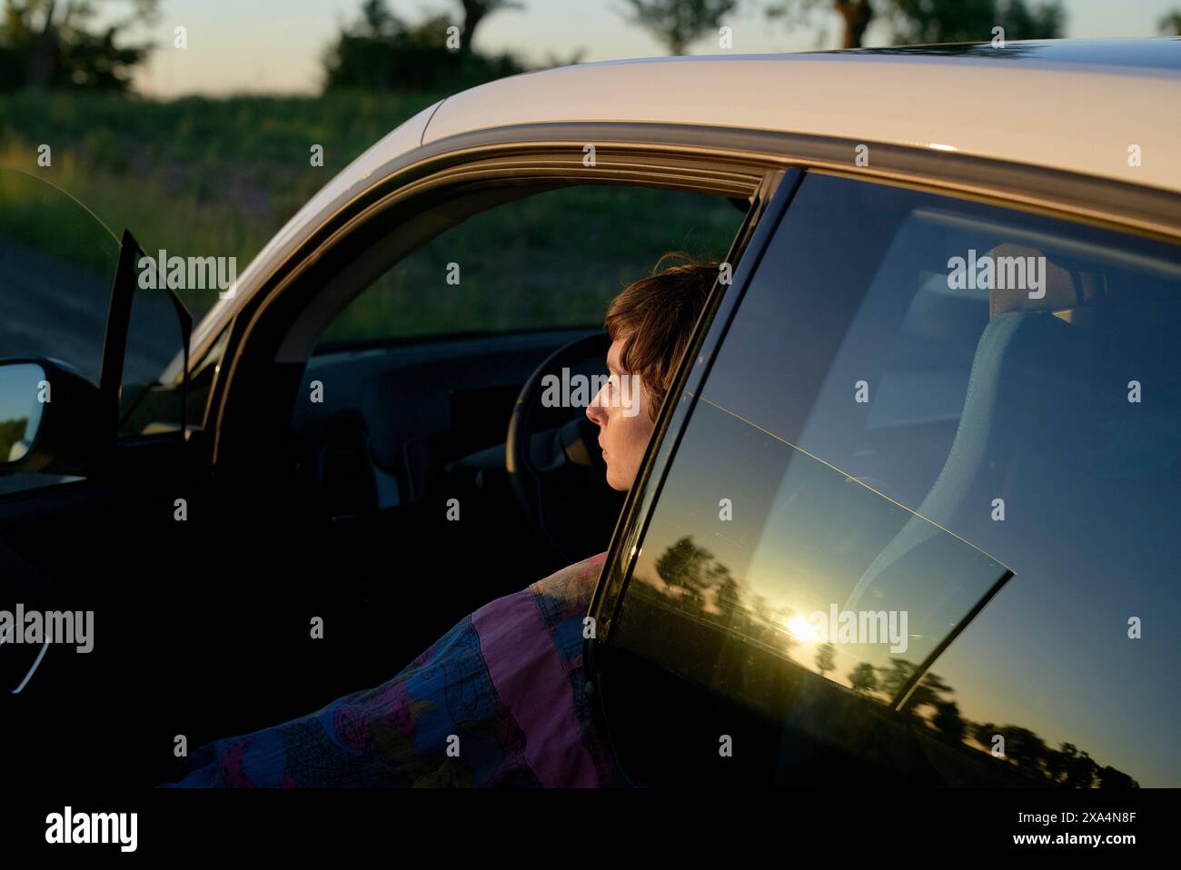 Une jeune femme est assise dans le siège du conducteur d'une voiture, avec leur silhouette visible à travers la fenêtre lorsque le soleil se couche en arrière-plan, projetant une lueur chaude. Banque D'Images