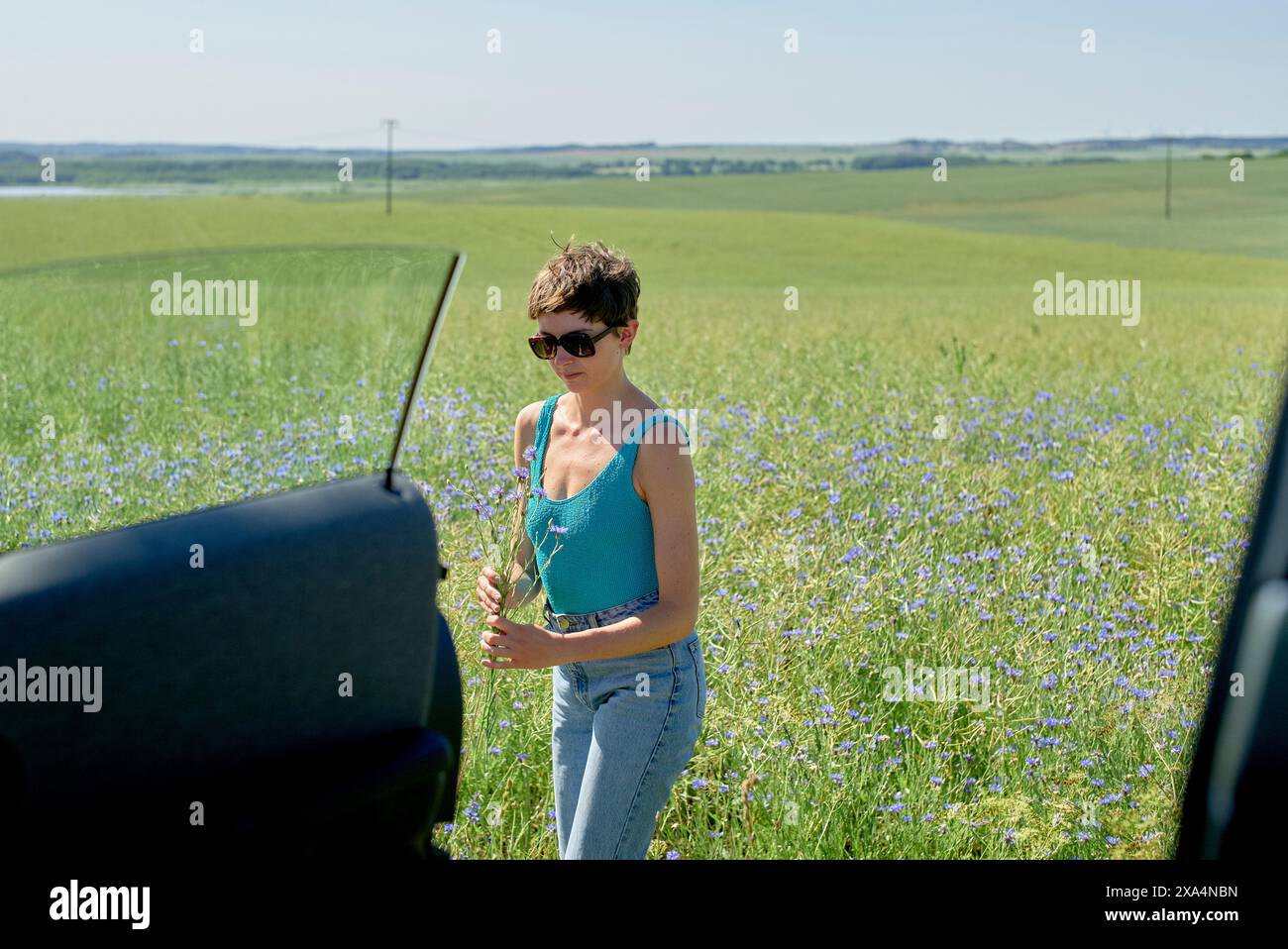 Une femme se tient à côté d'une voiture dans un champ d'herbe verte et de fleurs bleues par une journée ensoleillée, tenant un petit bouquet. Banque D'Images