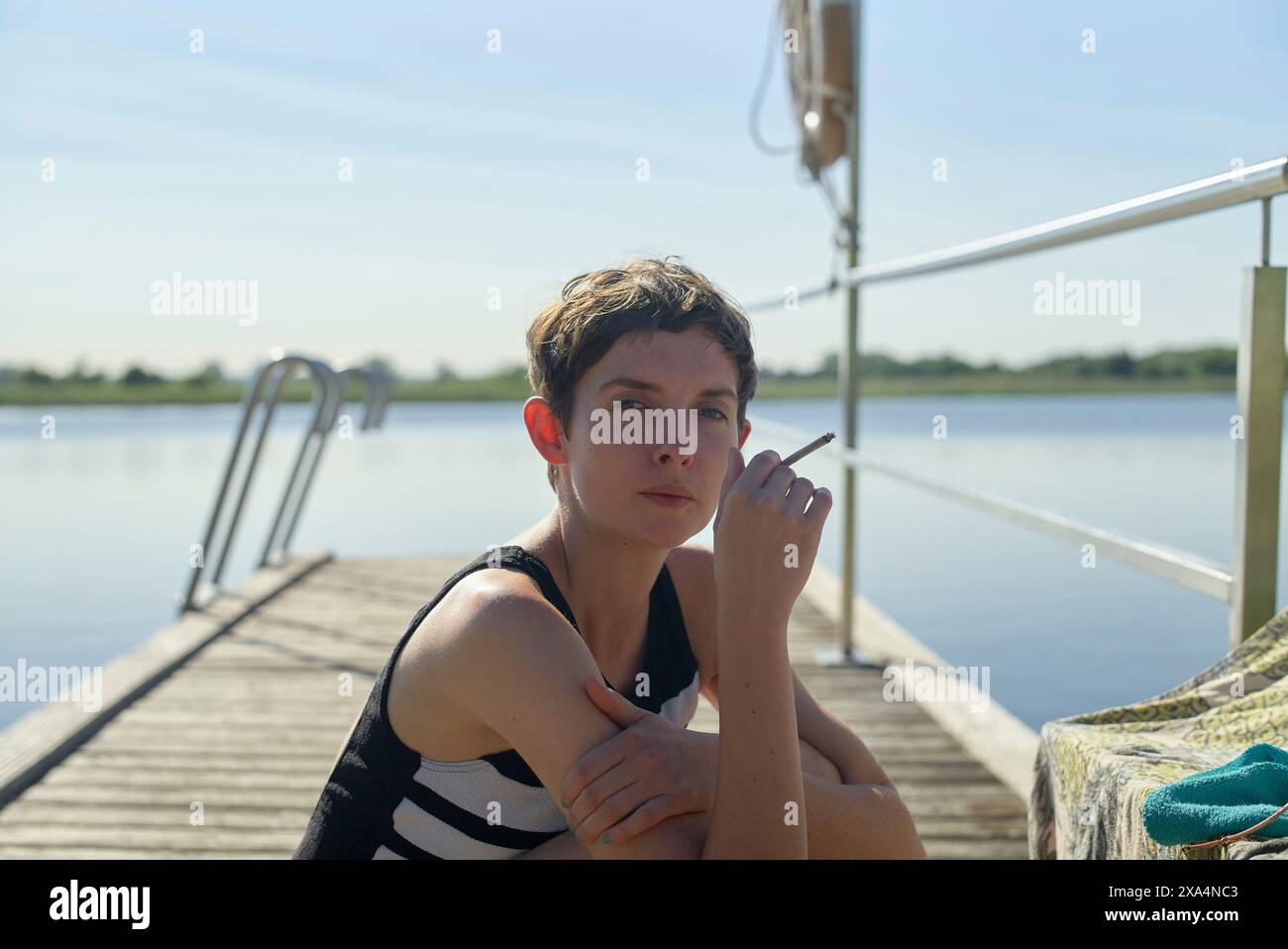 Une jeune femme est assise sur une jetée en bois près d'un lac, fumant une cigarette, avec une posture détendue sous un ciel bleu clair. Banque D'Images