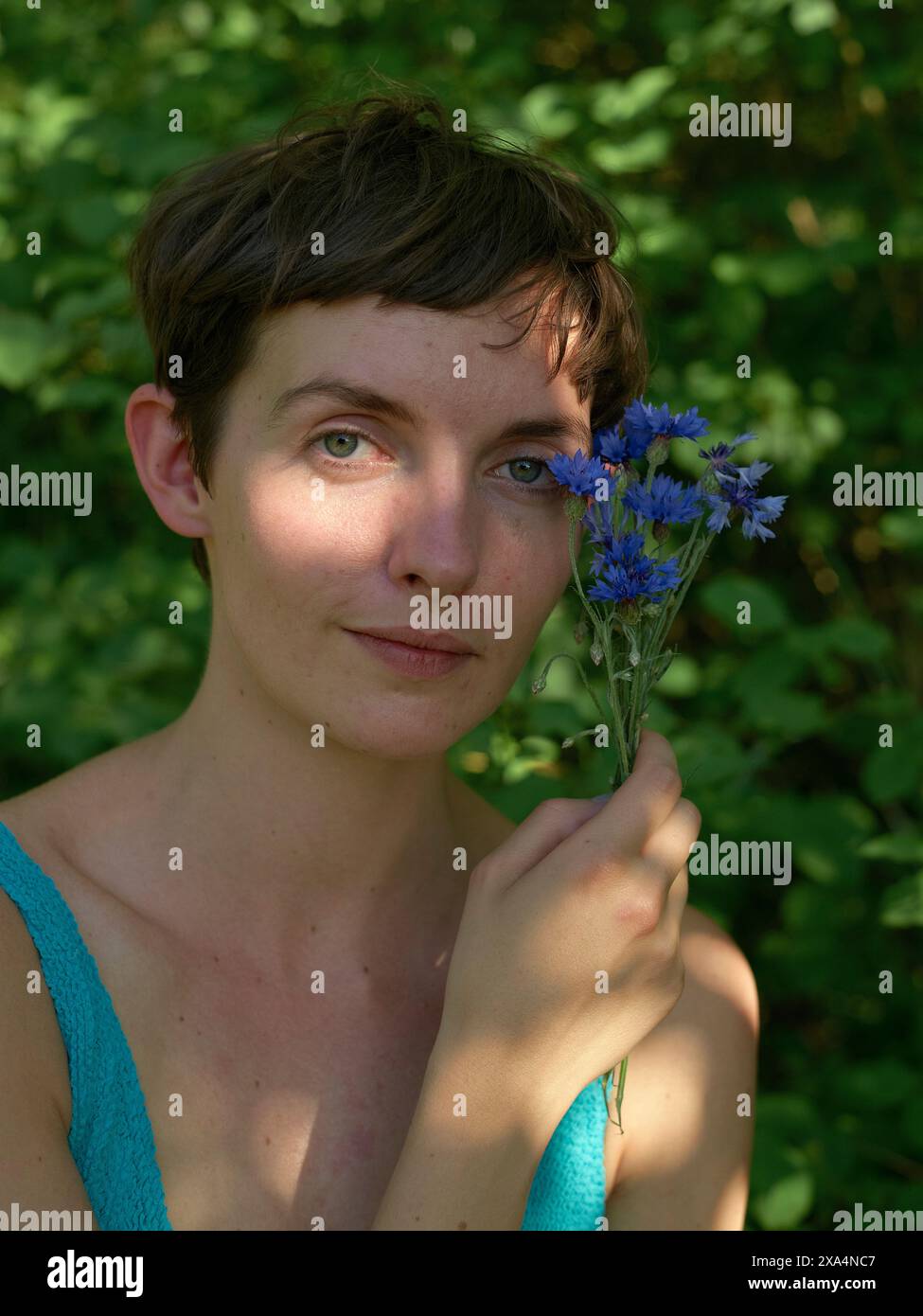 Une jeune femme aux cheveux courts tient un bouquet de fleurs bleues près de son visage, posant sur un fond vert naturel. Banque D'Images