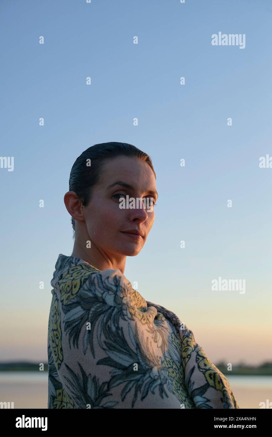 Une femme avec ses cheveux tirés en arrière se tient dans la lumière du soir, regardant par-dessus son épaule avec une expression calme contre un ciel clair. Banque D'Images