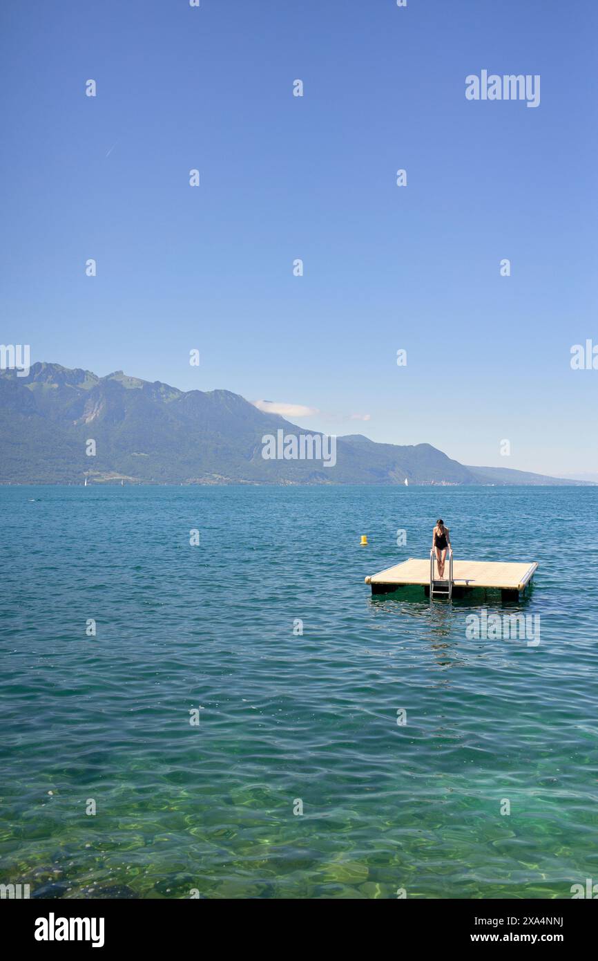 Une personne se tient sur un quai en bois qui s'étend dans un lac bleu clair, avec des montagnes et un ciel bleu en arrière-plan. Banque D'Images