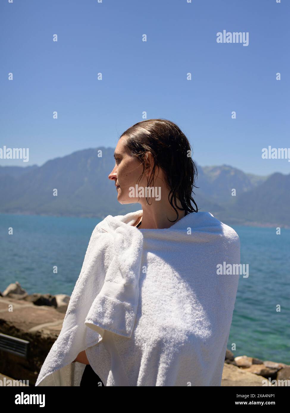 Une femme enveloppée dans une serviette blanche se tient au bord d'un lac, regardant au loin avec des montagnes et un ciel bleu clair en arrière-plan. Banque D'Images