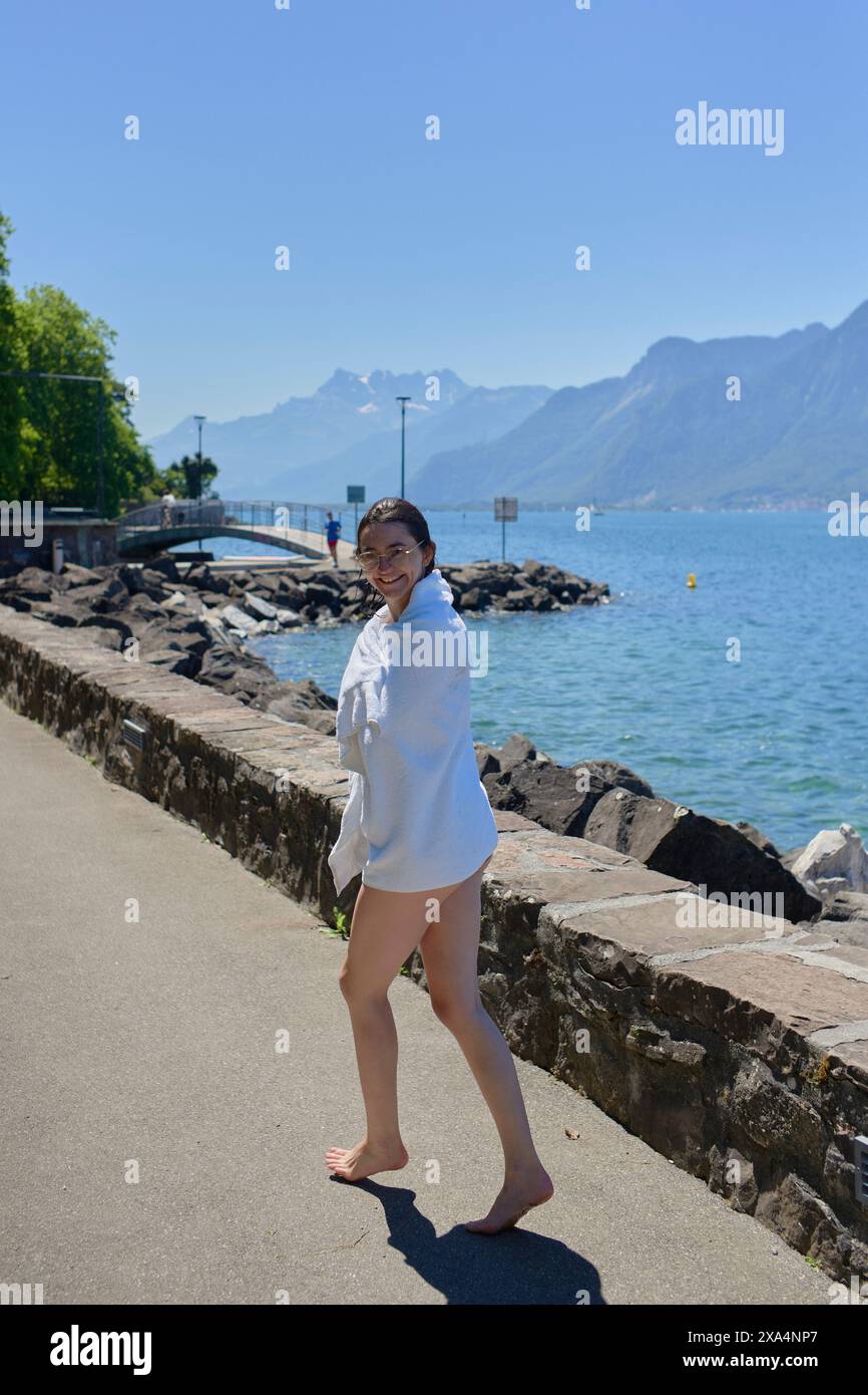 Une femme en chemise blanche marche pieds nus sur une promenade rocheuse au bord d'un lac pittoresque avec des montagnes en arrière-plan par une journée ensoleillée claire. Banque D'Images