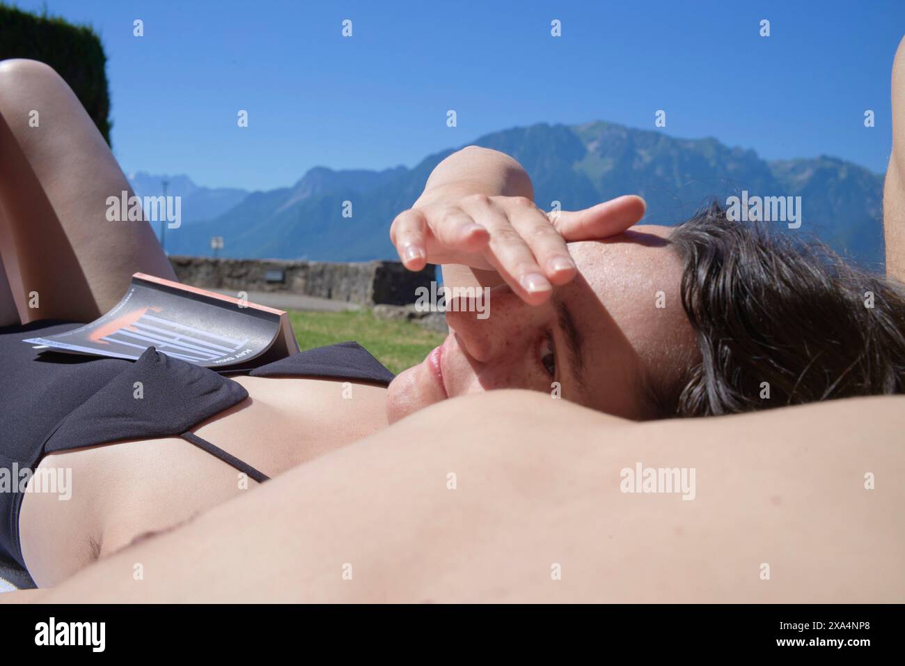 Une personne est allongée avec sa tête reposant sur son bras, protégeant ses yeux du soleil, avec des montagnes en arrière-plan. Banque D'Images