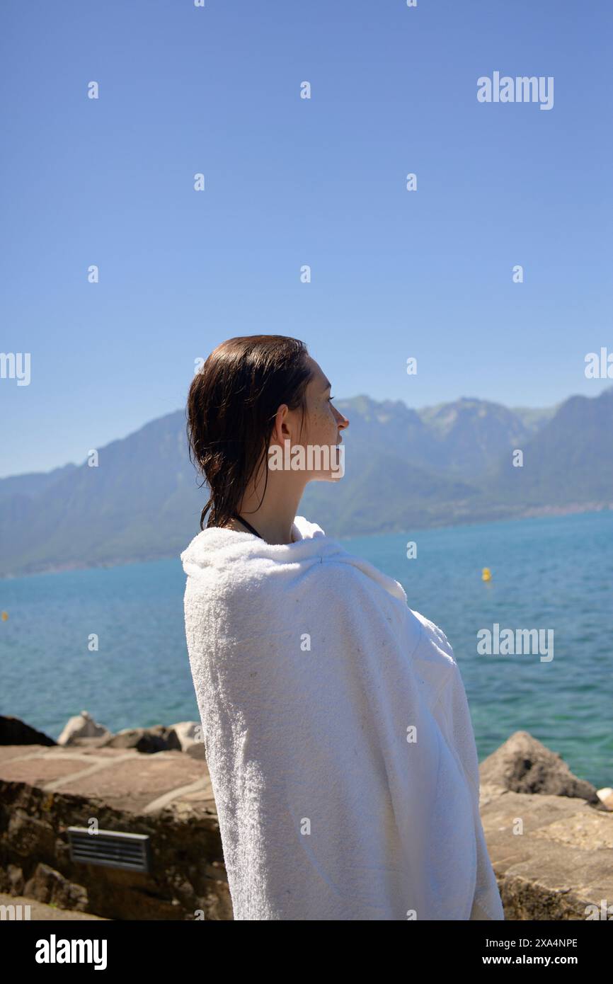 Une personne enveloppée dans une serviette blanche se tient près d'un lac regardant les montagnes sous un ciel bleu clair. Banque D'Images