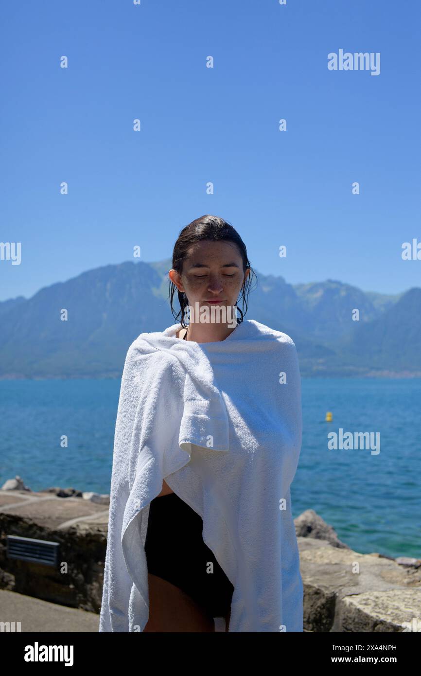 Une jeune femme se tient au bord d'un lac avec des montagnes en arrière-plan, drapée dans une serviette blanche et plissant légèrement à la lumière du soleil. Banque D'Images