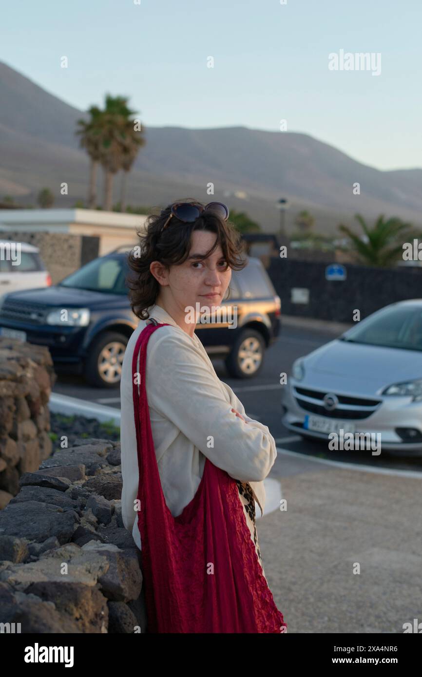 Une jeune femme souriante se tient dehors avec des voitures garées et une montagne en arrière-plan pendant ce qui semble être en fin d'après-midi. Banque D'Images