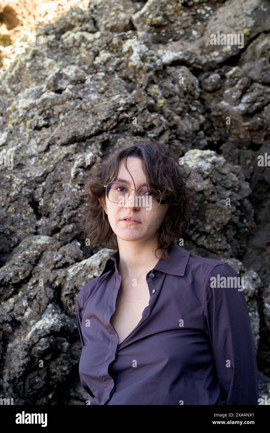 Une jeune femme aux cheveux bouclés courts et aux lunettes se tient devant un fond rocheux, portant une chemise boutonnée de couleur foncée. Banque D'Images