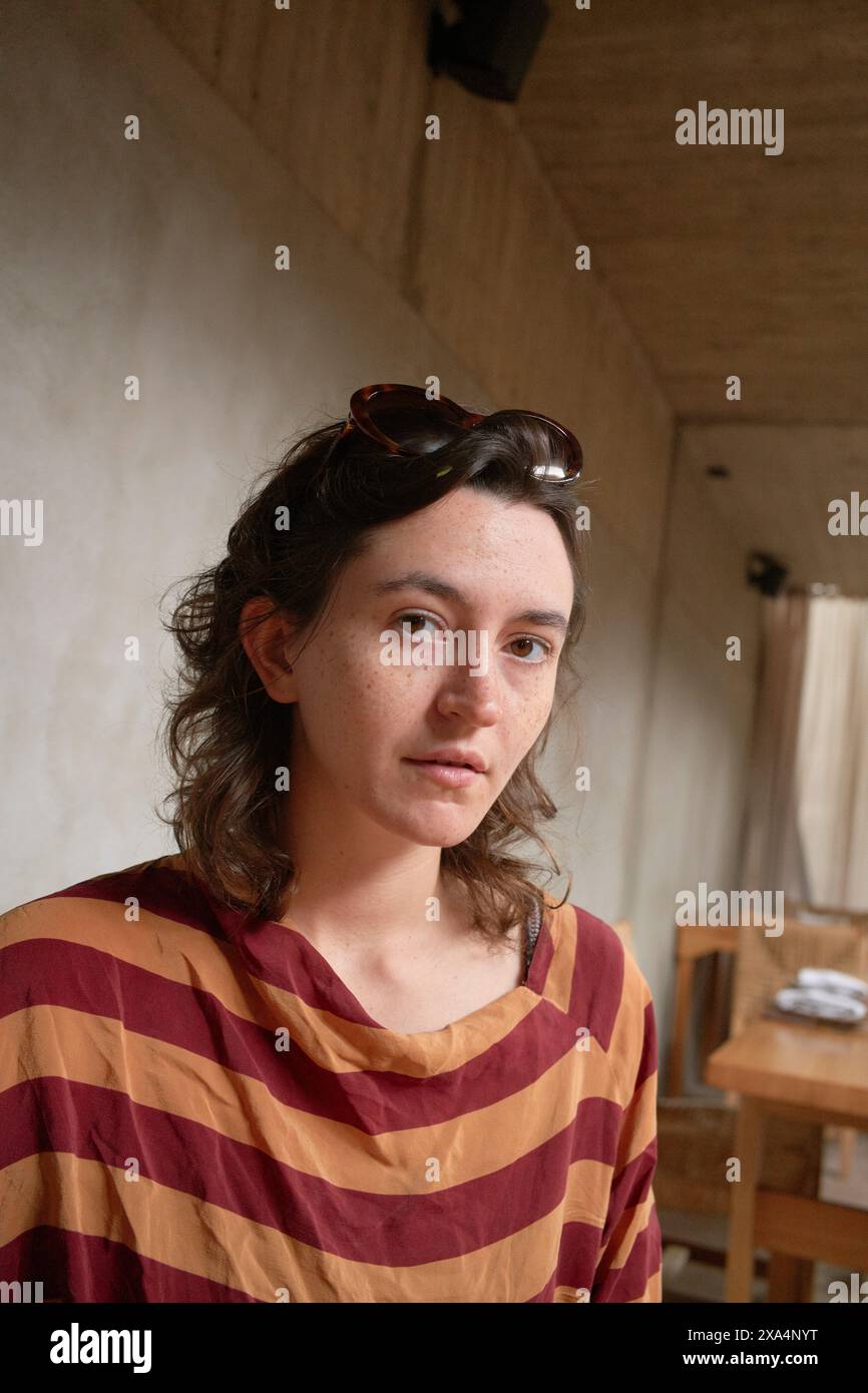 Une jeune femme avec des lunettes de soleil sur la tête porte une chemise rayée et regarde la caméra avec une expression neutre dans une pièce avec un éclairage doux. Banque D'Images